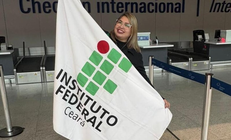 A imagem mostra a doutoranda Renata Teófilo de Sousa segurando a bandeira do IFCE no aeroporto de Fortaleza antes de embarcar para Macau, na China
